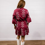 A woman viewed from behind wearing a burgundy mini dress with white floral embroidery, paired with white cowboy boots, standing against a white shiplap wall and wood floor.