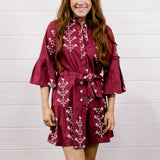 A woman modeling a short burgundy wrap dress with white floral embroidery and white cowboy boots, standing against a white wood-paneled wall and wood floor.