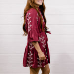 A woman modeling a short burgundy dress with white floral embroidery and three-quarter bell sleeves, paired with white cowboy boots, standing against a white wood-paneled wall and wooden floor.