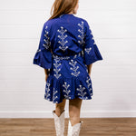 A woman viewed from behind wearing a royal blue kimono-style dress with white floral embroidery, paired with white cowboy boots, standing on a wooden floor against a white shiplap wall.