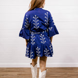 A woman viewed from behind wearing a royal blue kimono-style dress with white floral embroidery, paired with white cowboy boots, standing on a wooden floor against a white shiplap wall.
