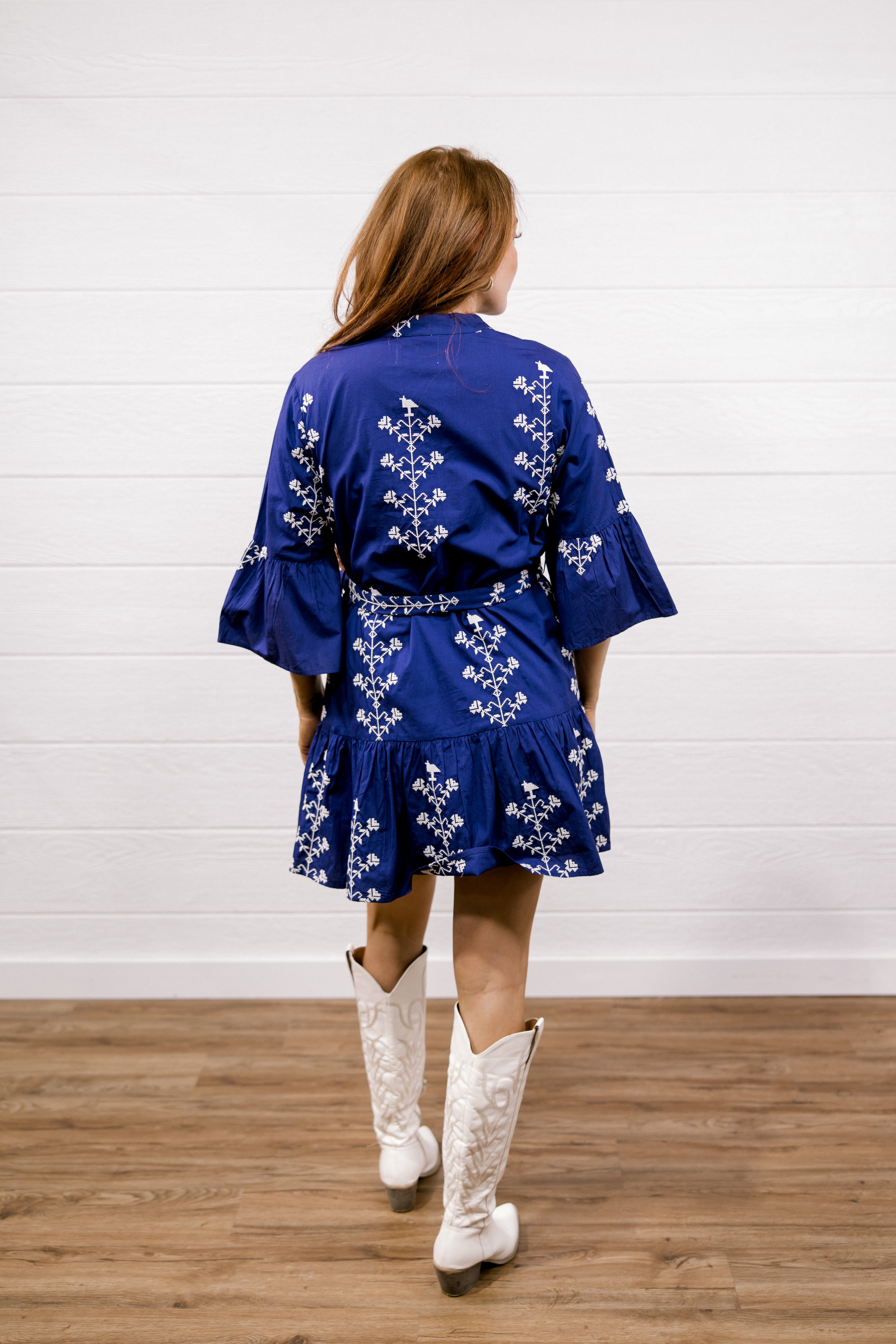 A woman viewed from behind wearing a royal blue kimono-style dress with white floral embroidery, paired with white cowboy boots, standing on a wooden floor against a white shiplap wall.