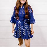 A woman modeling a purple wrap-style mini dress with white floral embroidery, paired with white cowboy boots, standing against a white shiplap wall and wood floor.