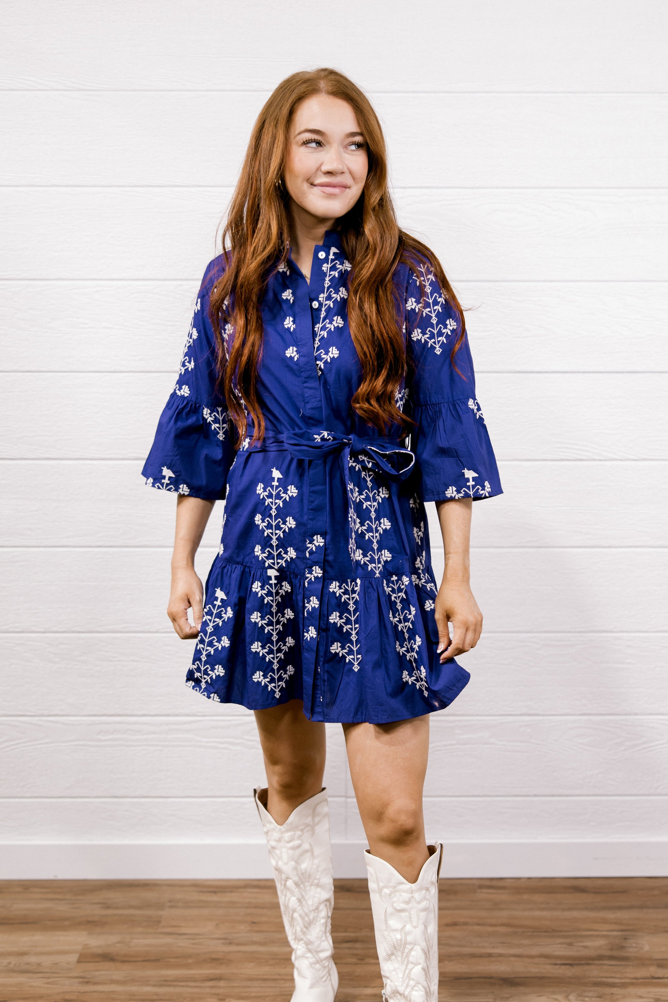 A woman modeling a purple wrap-style mini dress with white floral embroidery, paired with white cowboy boots, standing against a white shiplap wall and wood floor.