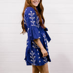 A young woman with long light brown hair is shown from the side and back, wearing a purple short dress with a white floral pattern and white cowboy boots, standing on a wooden floor against a white shiplap wall.