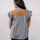 A woman viewed from behind wearing a gray and white vertically striped blouse with a rust-orange patterned yoke and ruffled cap sleeves, paired with dark jeans, set against a white paneled wall.