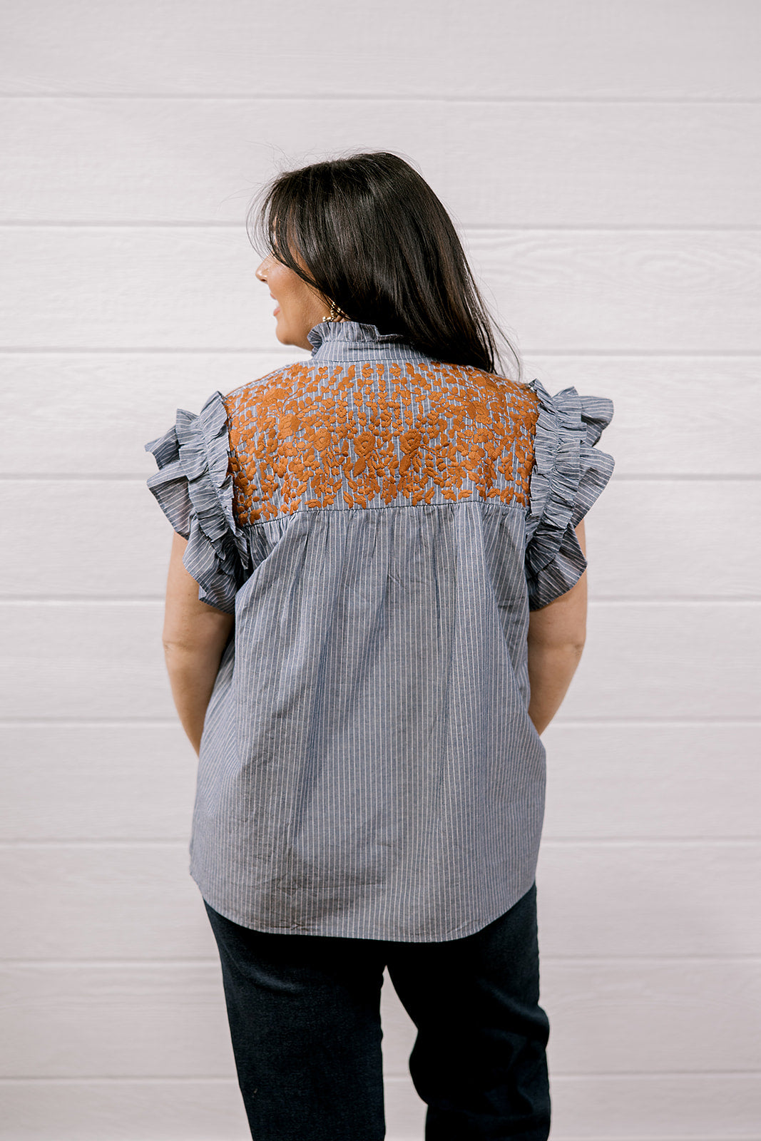 A woman viewed from behind wearing a gray and white vertically striped blouse with a rust-orange patterned yoke and ruffled cap sleeves, paired with dark jeans, set against a white paneled wall.