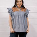 A woman modeling a sleeveless top with ruffled flutter sleeves, featuring a blue and white vertical stripe body and a rust-orange floral yoke across the neckline, paired with dark jeans against a white wood-paneled wall.