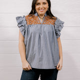 A woman modeling a sleeveless top with ruffled flutter sleeves, featuring a blue and white vertical stripe body and a rust-orange floral yoke across the neckline, paired with dark jeans against a white wood-paneled wall.