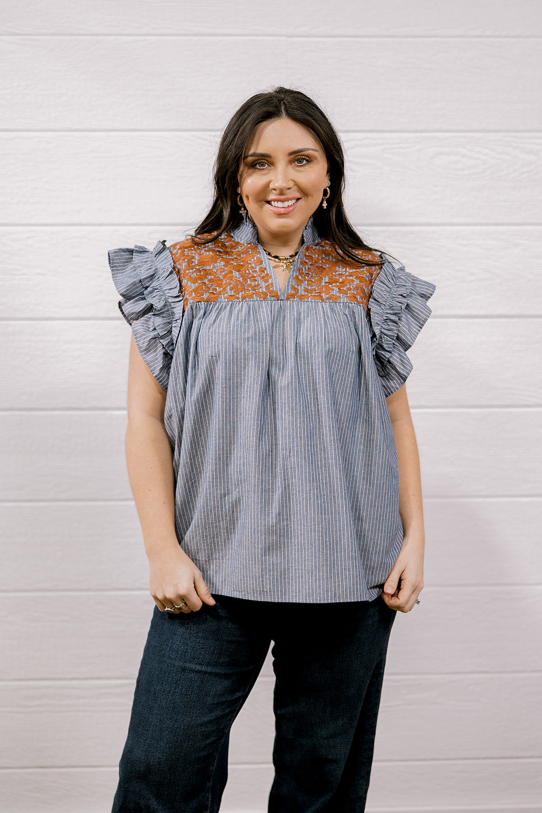 A woman modeling a sleeveless top with ruffled flutter sleeves, featuring a blue and white vertical stripe body and a rust-orange floral yoke across the neckline, paired with dark jeans against a white wood-paneled wall.