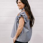A woman with long dark hair is shown from the side, wearing a sleeveless ruffled blouse with vertical blue and white stripes and orange-brown embroidered detailing on the shoulders, paired with dark jeans, against a white wood-paneled wall.