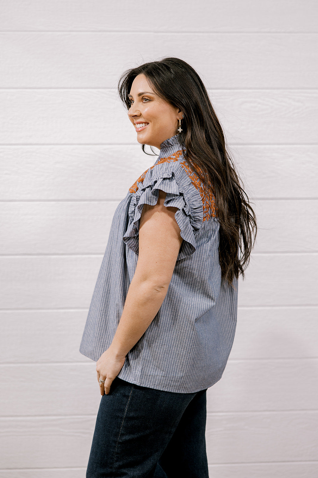 A woman with long dark hair is shown from the side, wearing a sleeveless ruffled blouse with vertical blue and white stripes and orange-brown embroidered detailing on the shoulders, paired with dark jeans, against a white wood-paneled wall.