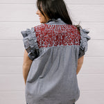 A woman shown from the back wearing a sleeveless blouse with ruffled short sleeves, a gray and white pinstripe body, and a red floral embroidered yoke panel, set against a white paneled wall.