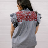 A woman shown from the back wearing a sleeveless blouse with ruffled short sleeves, a gray and white pinstripe body, and a red floral embroidered yoke panel, set against a white paneled wall.