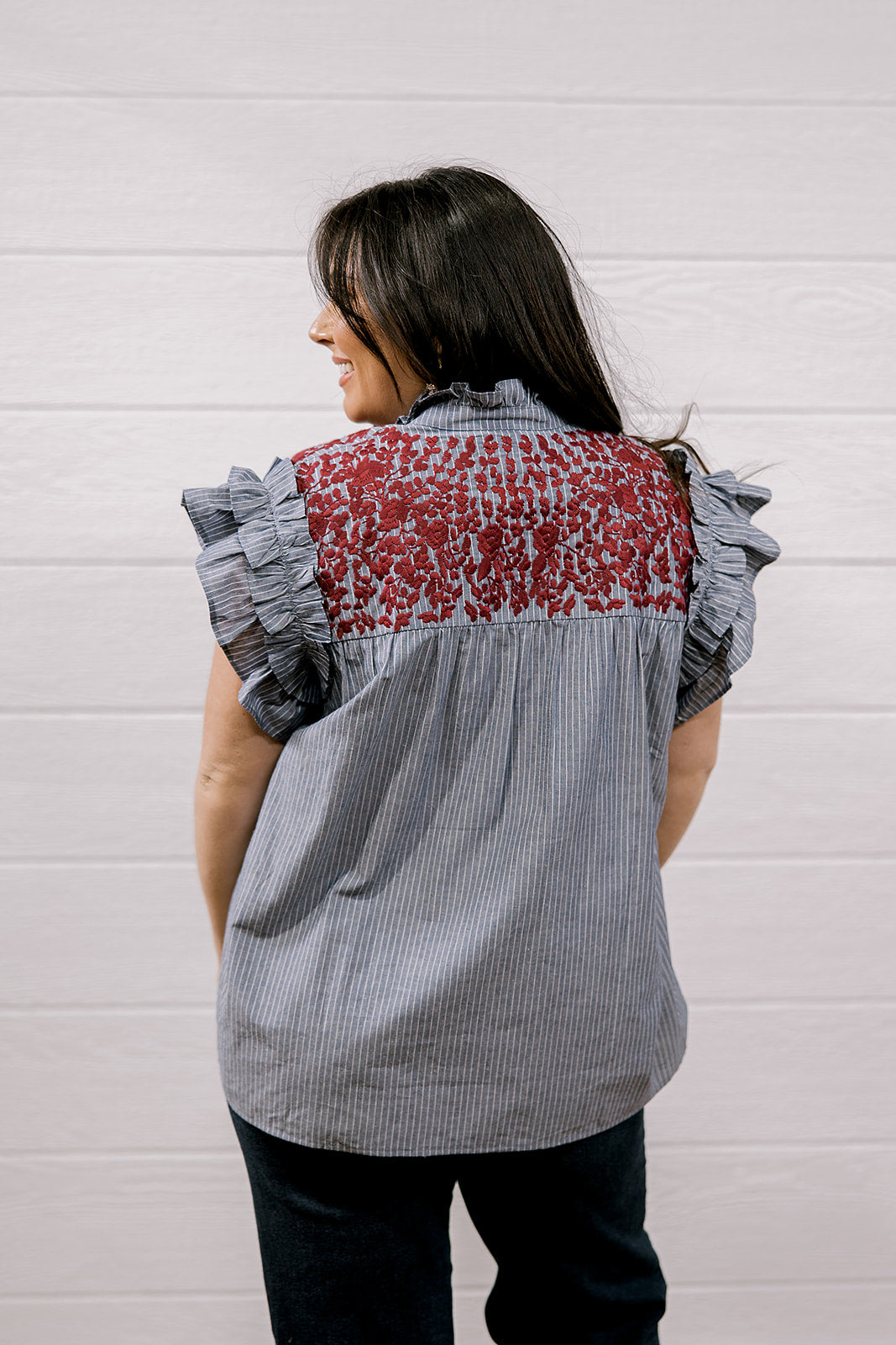A woman shown from the back wearing a sleeveless blouse with ruffled short sleeves, a gray and white pinstripe body, and a red floral embroidered yoke panel, set against a white paneled wall.