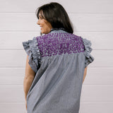 A woman shown from the back wearing a gray and white vertically striped blouse with a purple patterned yoke and ruffled cap sleeves, set against a white wood paneled wall.