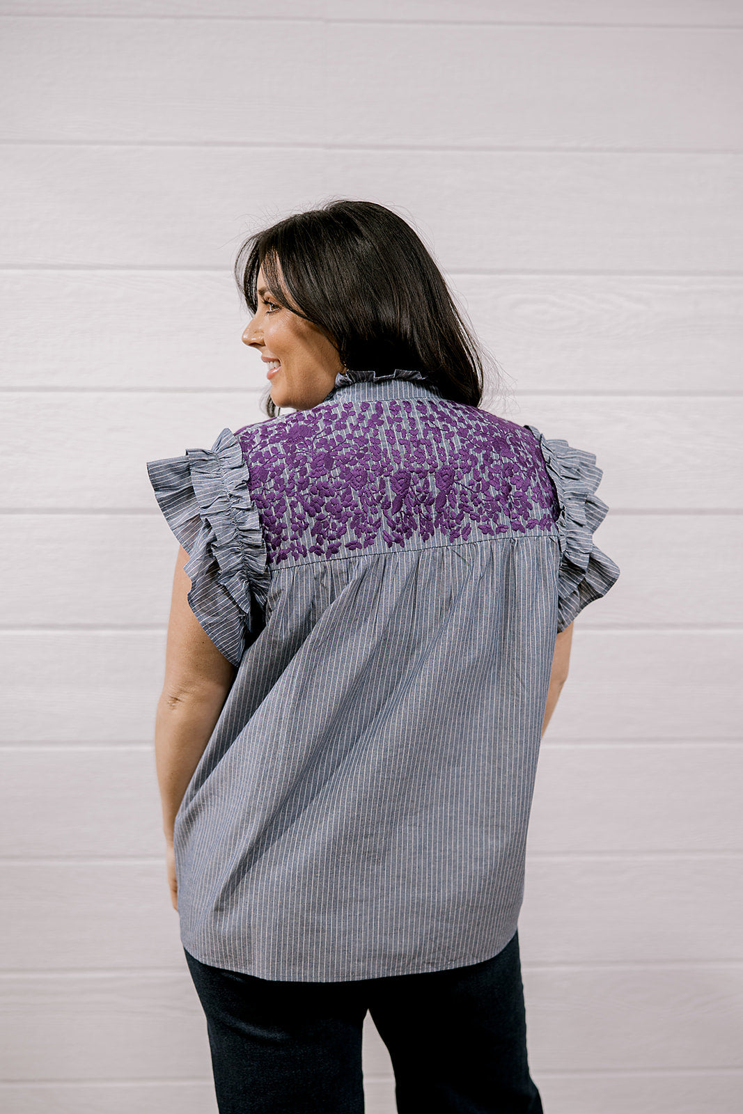 A woman shown from the back wearing a gray and white vertically striped blouse with a purple patterned yoke and ruffled cap sleeves, set against a white wood paneled wall.