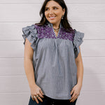 A woman modeling a sleeveless top with ruffled cap sleeves, featuring a gray and white vertical stripe body and a purple floral yoke, paired with dark jeans against a white wood-paneled wall.