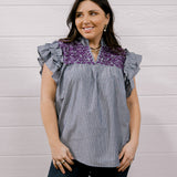 A woman modeling a sleeveless top with ruffled cap sleeves, featuring a gray and white vertical stripe body and a purple floral yoke, paired with dark jeans against a white wood-paneled wall.