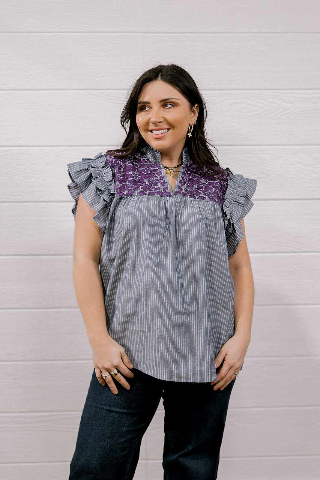 A woman modeling a sleeveless top with ruffled cap sleeves, featuring a gray and white vertical stripe body and a purple floral yoke, paired with dark jeans against a white wood-paneled wall.