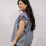 A woman wearing a sleeveless ruffled blouse with vertical gray and white pinstripes and purple lace trim at the neckline, paired with dark jeans, standing against a white paneled wall.