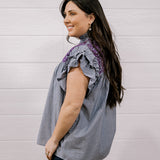 A woman wearing a sleeveless ruffled blouse with vertical gray and white pinstripes and purple lace trim at the neckline, paired with dark jeans, standing against a white paneled wall.
