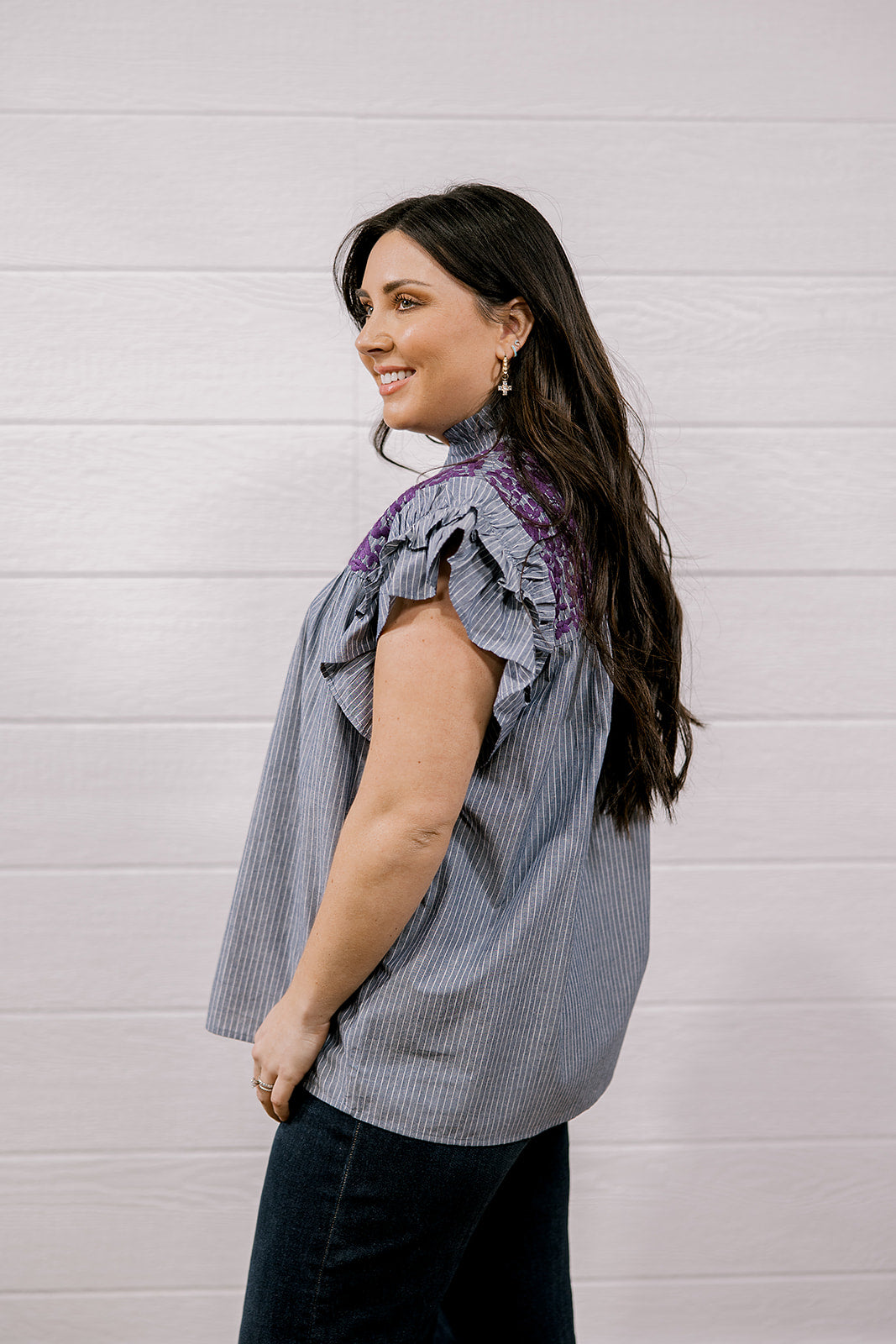 A woman wearing a sleeveless ruffled blouse with vertical gray and white pinstripes and purple lace trim at the neckline, paired with dark jeans, standing against a white paneled wall.