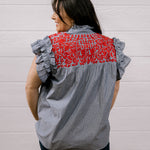 A woman shown from the back wearing a gray and white vertically striped blouse with ruffled sleeves and a red floral embroidered yoke, paired with dark pants, set against a white paneled wall.
