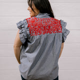A woman shown from the back wearing a gray and white vertically striped blouse with ruffled sleeves and a red floral embroidered yoke, paired with dark pants, set against a white paneled wall.