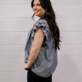 A woman with long dark hair is shown from the side, wearing a sleeveless ruffled blouse with a blue and white vertical stripe pattern and red accents, paired with dark pants, against a white paneled wall.