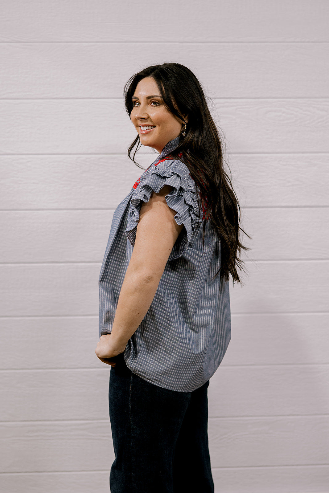 A woman with long dark hair is shown from the side, wearing a sleeveless ruffled blouse with a blue and white vertical stripe pattern and red accents, paired with dark pants, against a white paneled wall.