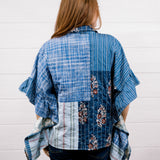 A woman viewed from behind wearing a blue patchwork shirt with plaid, striped, and floral patterns, paired with dark blue jeans, standing against a white wood-paneled wall.