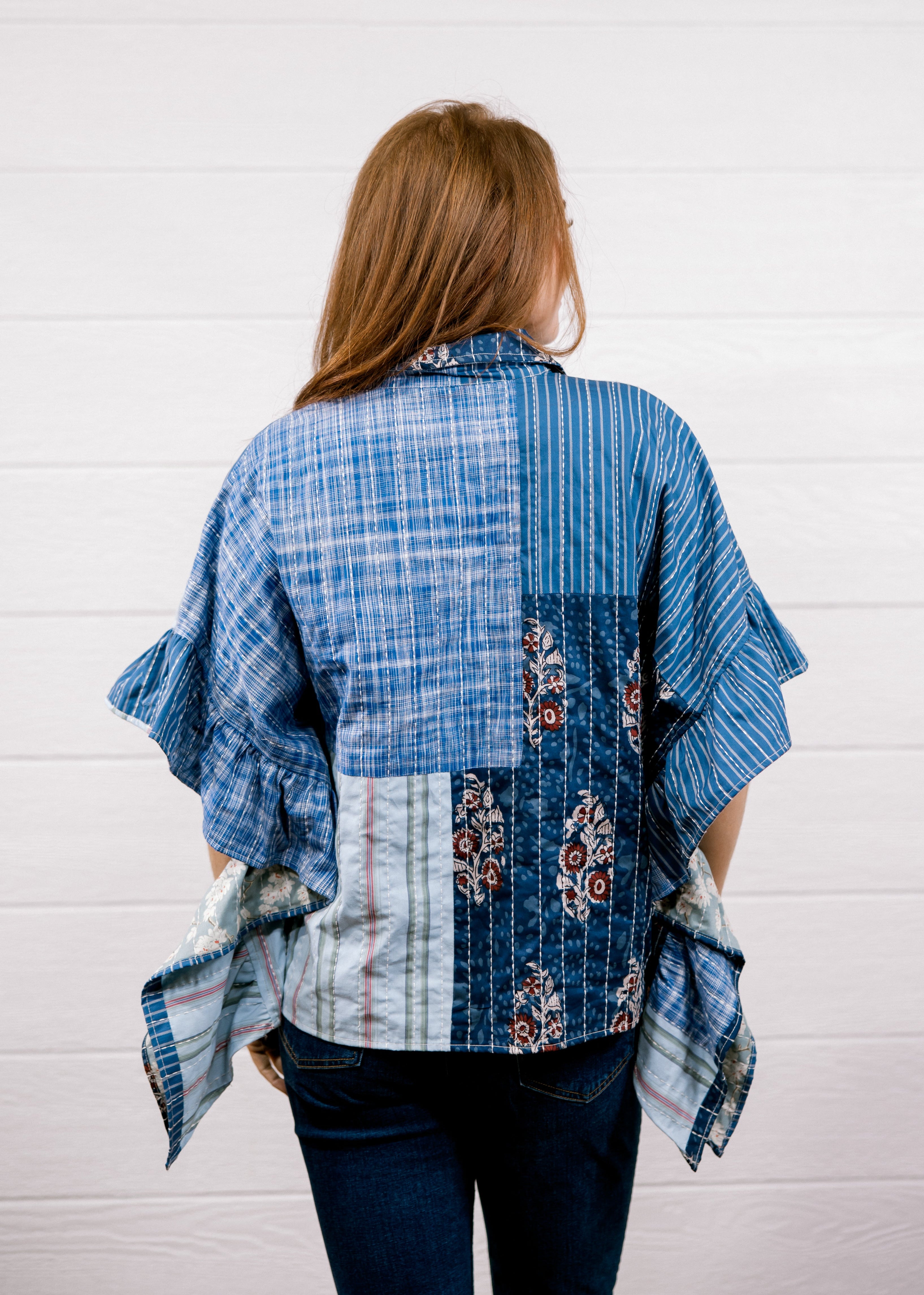 A woman viewed from behind wearing a blue patchwork shirt with plaid, striped, and floral patterns, paired with dark blue jeans, standing against a white wood-paneled wall.