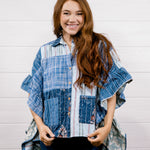 A smiling woman with long wavy brown hair stands against a white paneled wall wearing a blue and white patchwork button-up shirt with ruffled sleeves and dark blue jeans.