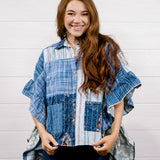 A smiling woman with long wavy brown hair stands against a white paneled wall wearing a blue and white patchwork button-up shirt with ruffled sleeves and dark blue jeans.