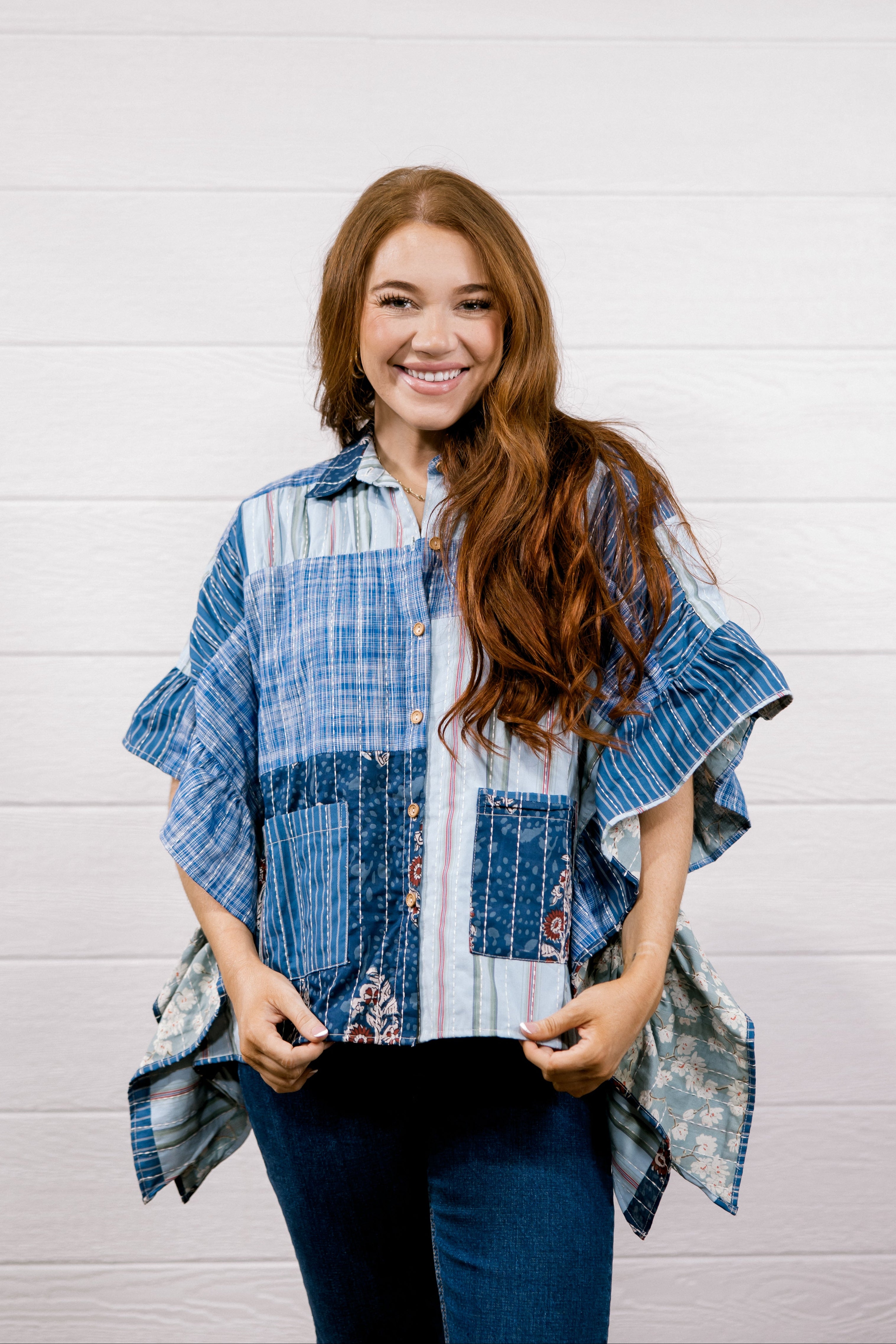 A smiling woman with long wavy brown hair stands against a white paneled wall wearing a blue and white patchwork button-up shirt with ruffled sleeves and dark blue jeans.