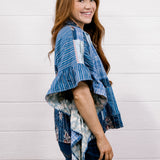 A woman with long light brown hair is shown from the side, smiling, wearing a blue and white patterned blouse with ruffled sleeves and dark blue jeans, standing against a white wood-paneled wall.