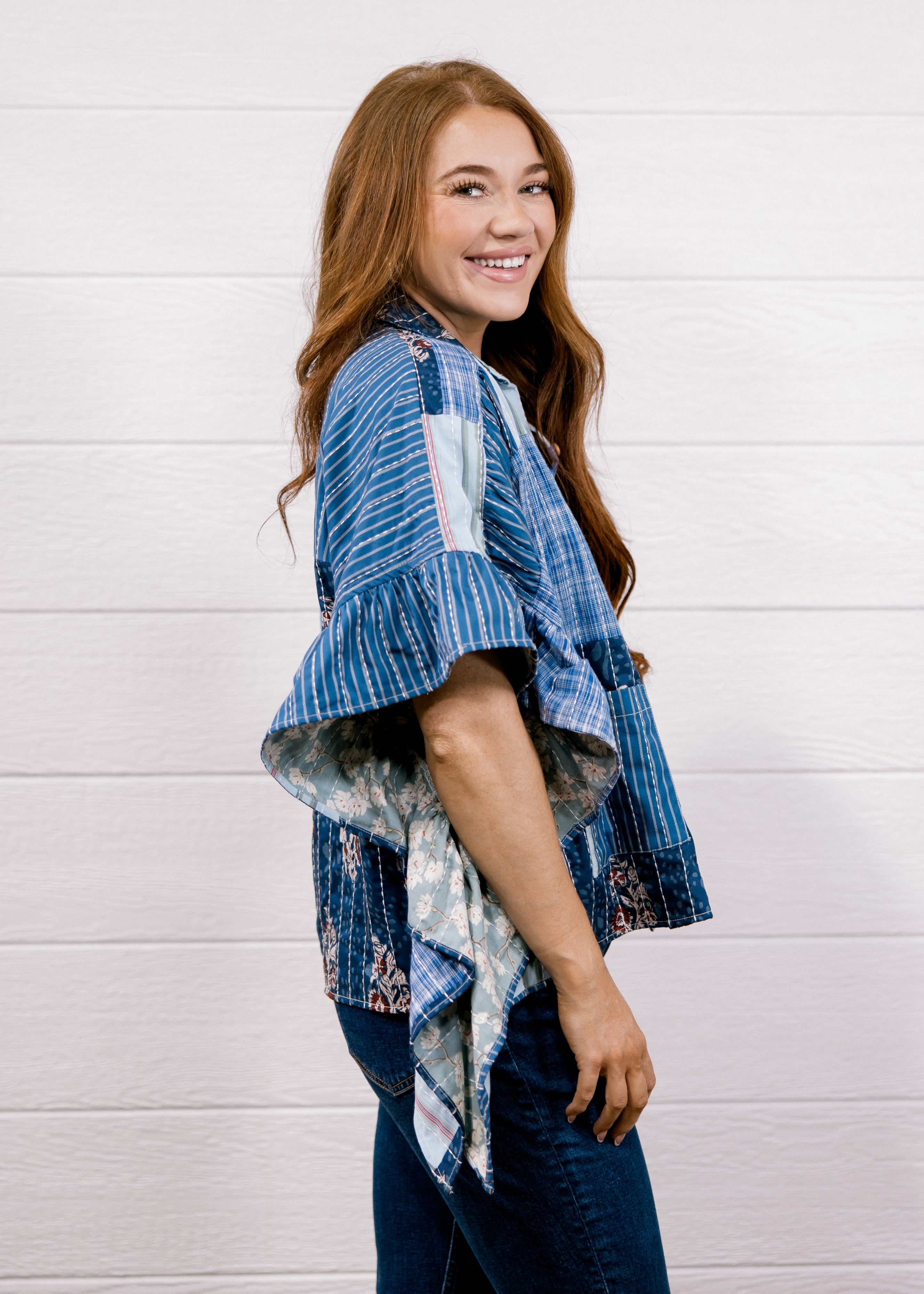 A woman with long light brown hair is shown from the side, smiling, wearing a blue and white patterned blouse with ruffled sleeves and dark blue jeans, standing against a white wood-paneled wall.