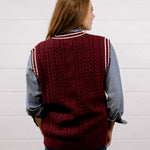 A woman viewed from behind wearing a burgundy cable-knit sweater vest with white striped ribbed trim over a blue and white striped button-down shirt and blue jeans, standing against a white wood-paneled wall.