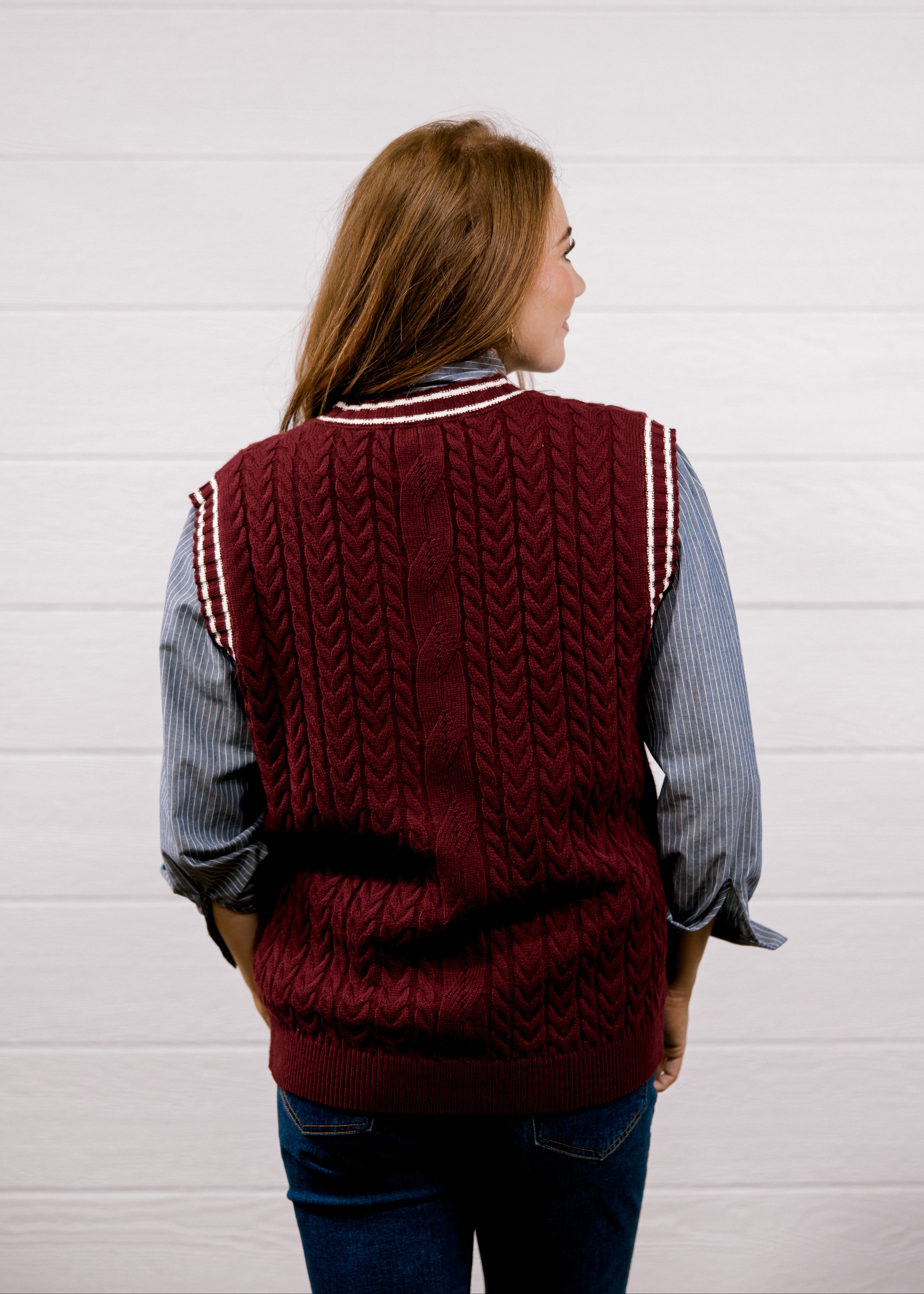 A woman viewed from behind wearing a burgundy cable-knit sweater vest with white striped ribbed trim over a blue and white striped button-down shirt and blue jeans, standing against a white wood-paneled wall.