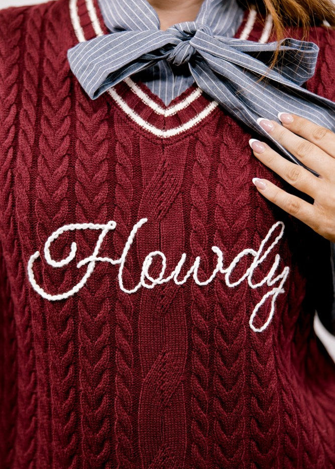 A close-up of a burgundy cable-knit sweater vest with white embroidered script text reading 'Howdy' across the chest, layered over a gray and white pinstriped collared shirt with a large tied bow at the neck.