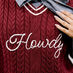 A close-up of a burgundy cable-knit sweater vest with white embroidered script text reading 'Howdy' across the chest, layered over a gray and white pinstriped collared shirt with a large tied bow at the neck.