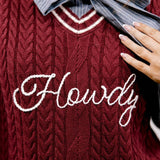 A close-up of a burgundy cable-knit sweater vest with white embroidered script text reading 'Howdy' across the chest, layered over a gray and white pinstriped collared shirt with a large tied bow at the neck.