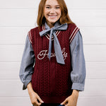 A smiling young woman stands against a white wood-plank wall wearing a burgundy cable-knit sweater vest over a blue-and-white striped long-sleeve shirt with a matching blue bow tie, paired with blue jeans.