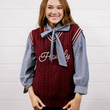A smiling young woman stands against a white wood-plank wall wearing a burgundy cable-knit sweater vest over a blue-and-white striped long-sleeve shirt with a matching blue bow tie, paired with blue jeans.