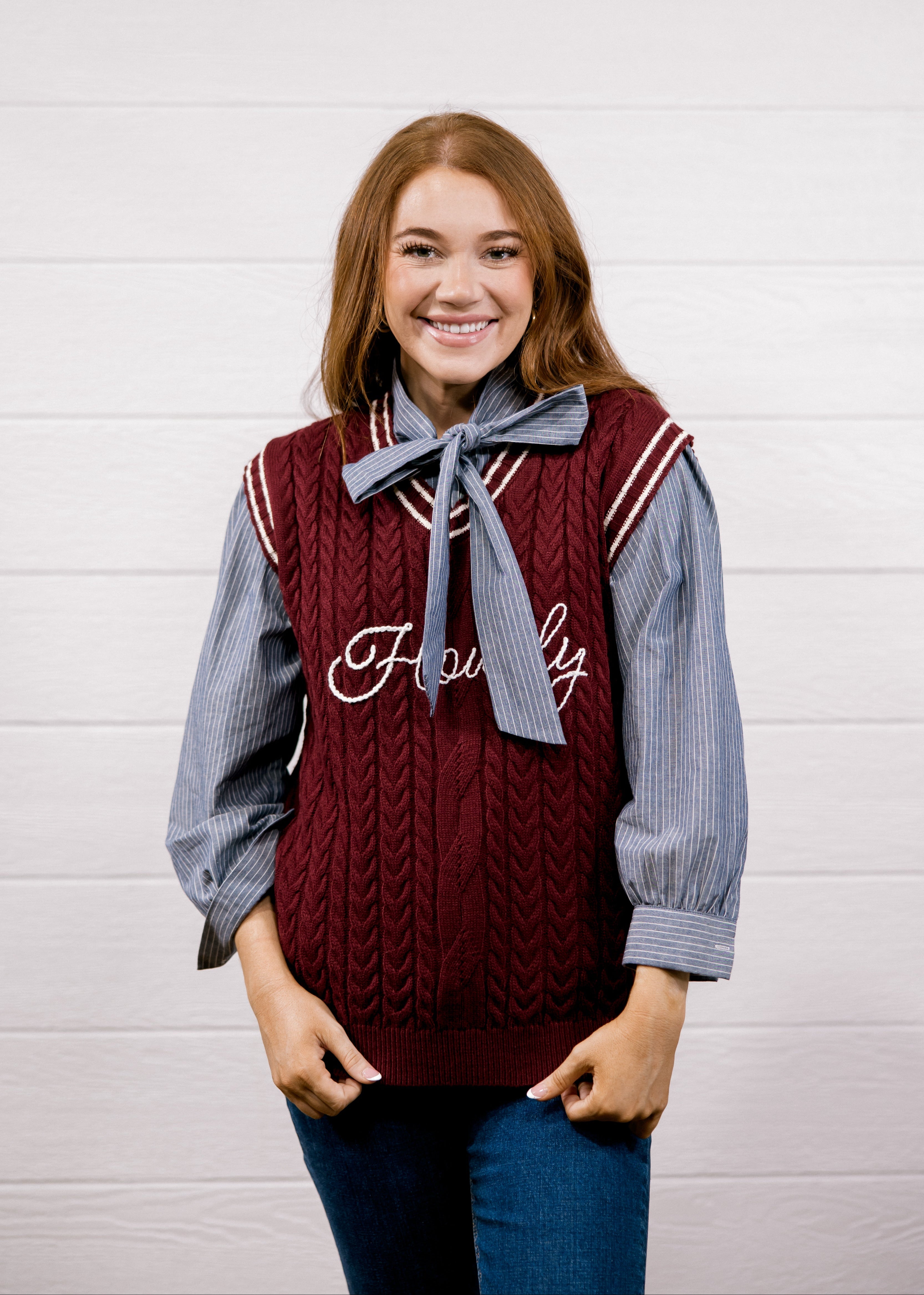 A smiling young woman stands against a white wood-plank wall wearing a burgundy cable-knit sweater vest over a blue-and-white striped long-sleeve shirt with a matching blue bow tie, paired with blue jeans.