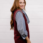 A young woman with long light brown hair is shown from the side and back, wearing a burgundy cable-knit sweater vest over a blue and white striped button-down shirt, paired with dark blue jeans. She is standing against a white wood-paneled wall.
