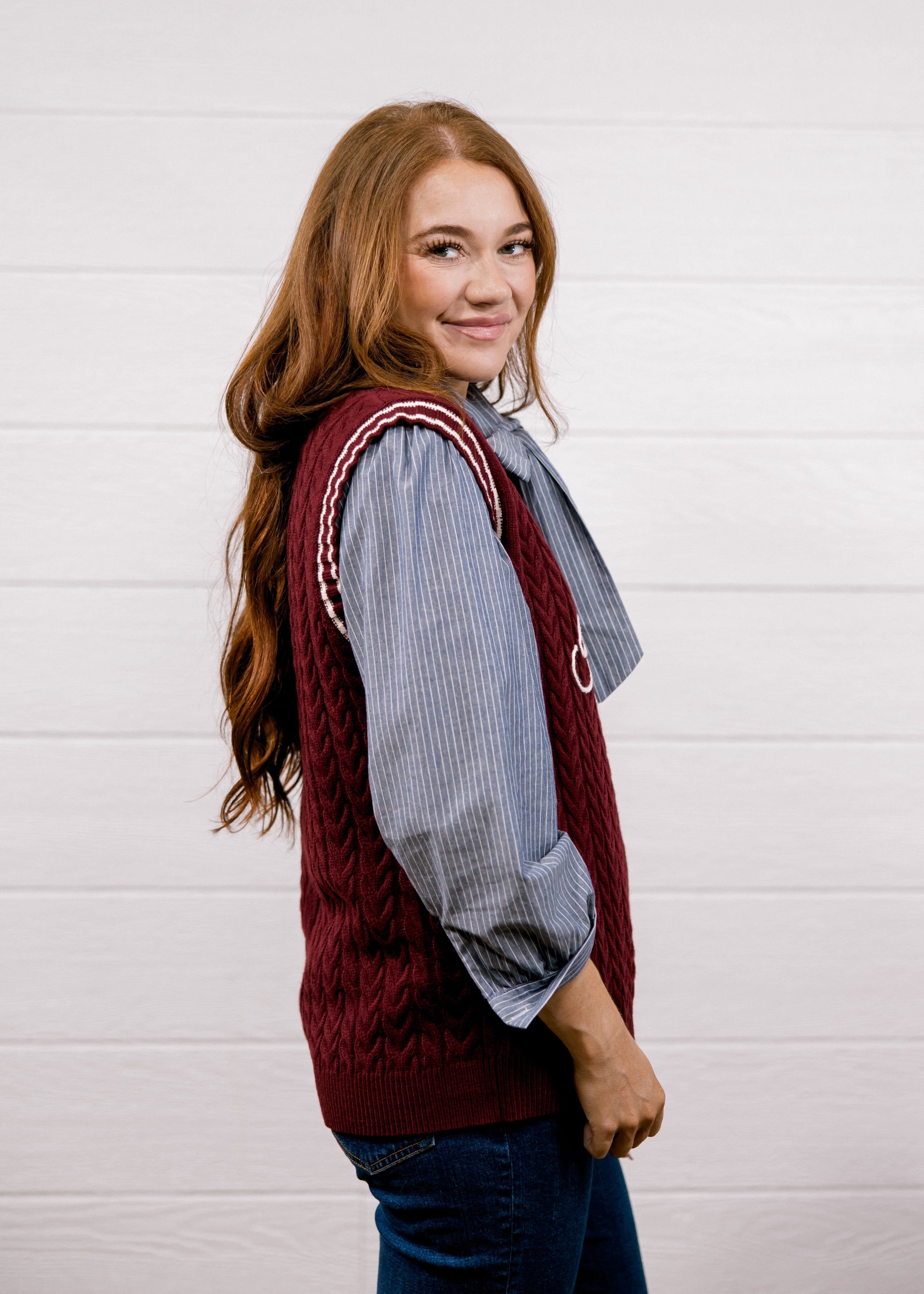 A young woman with long light brown hair is shown from the side and back, wearing a burgundy cable-knit sweater vest over a blue and white striped button-down shirt, paired with dark blue jeans. She is standing against a white wood-paneled wall.