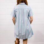A woman viewed from behind wearing a light blue short-sleeve striped shirt dress with thin red and white pinstripes, paired with brown cowboy boots, standing against a white paneled wall on a wood floor.