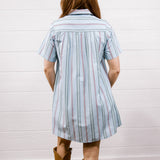 A woman viewed from behind wearing a light blue short-sleeve striped shirt dress with thin red and white pinstripes, paired with brown cowboy boots, standing against a white paneled wall on a wood floor.